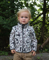 Child wearing a patterned jacket standing outdoors with greenery in the background