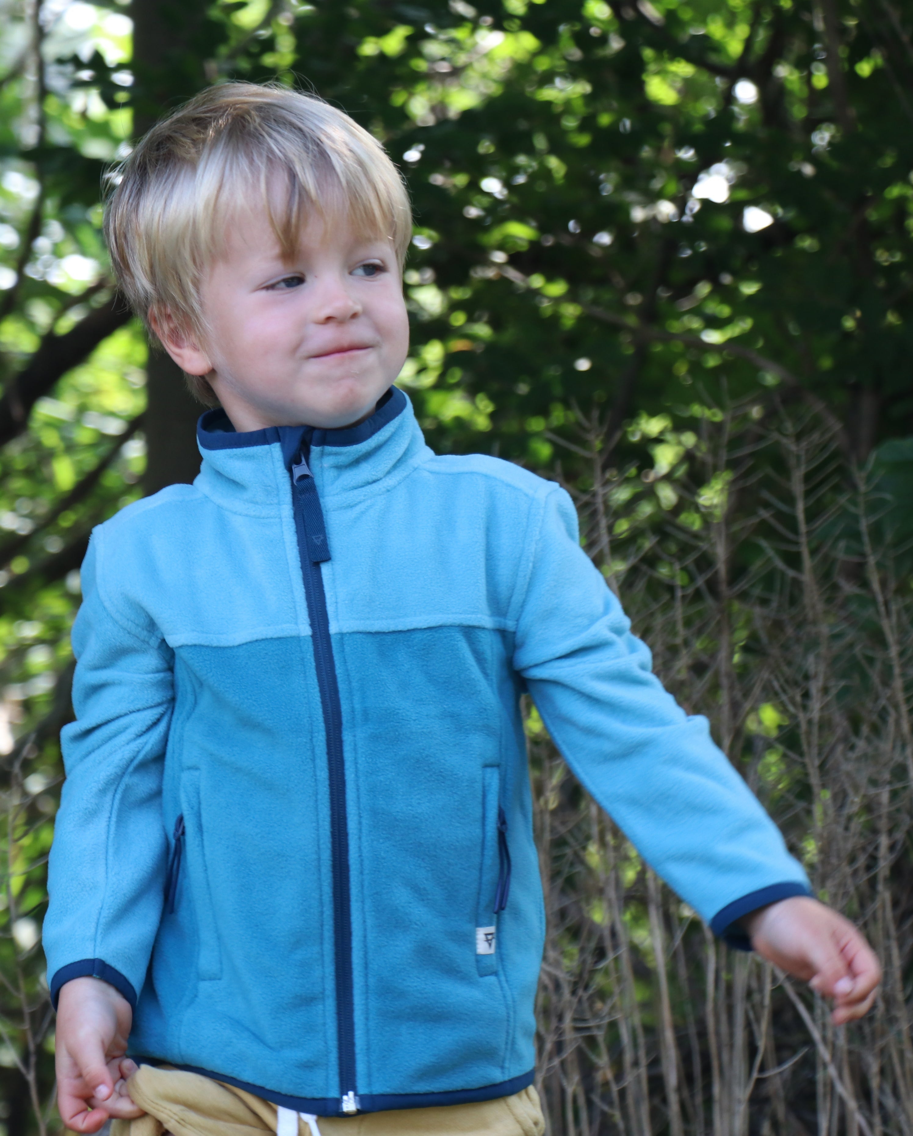 Child wearing a blue jacket standing outdoors with greenery in the background